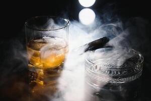 A glass of whiskey or Bourbon with ice cubes and a cigar on a black slate with smoke photo