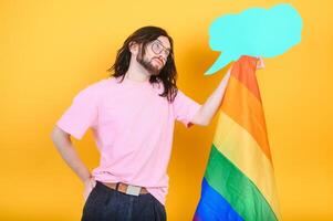 Portrait of cheerful smiling homosexual man holding empty blank board, with a gay pride flag at studio. Lgbtq flag, rainbow flag, celebrating parade. photo