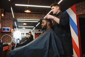 retrato de joven negro hombre siendo recortado con profesional eléctrico clíper máquina en barberia.masculino belleza tratamiento concepto. joven africano chico consiguiendo nuevo Corte de pelo en Barbero salón foto