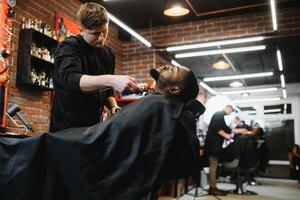 un Barbero es yendo mediante el eléctrico corte y afeitado máquina para el barba de un afroamericano brasileño chico. foto