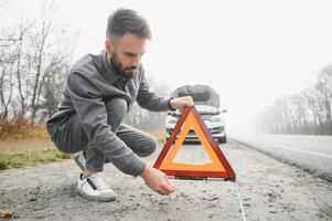 un joven hombre cerca un roto coche con un abierto capucha en el borde del camino. foto