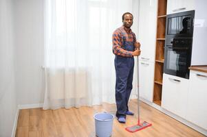 profesional limpiador en azul uniforme Lavado piso y limpiando polvo desde el mueble en el vivo habitación de el departamento. limpieza Servicio concepto foto