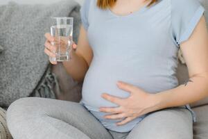 Pregnant woman with glass of water sitting on sofa. Young expectant blonde rest at home, copy space. Pregnancy, healthcare, thirst concept photo