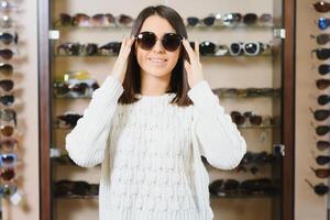 Which one is better fit me. Portrait of cheerful hesitating woman in optician store, making decision, holding stylish sunglasses, choosing what she should buy. Great discounts for buying two pairs. photo