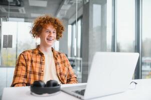 Young happy male freelancer in casual clothes sitting in cafe with laptop and using mobile phone. photo