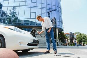 joven hermoso hombre participación cargando cable a eléctrico cargando estación punto en pie cerca su nuevo coche , mirando satisfecho. foto