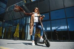 Young beautiful woman in a jacket smiles and rides an electric scooter to work along office buildings photo