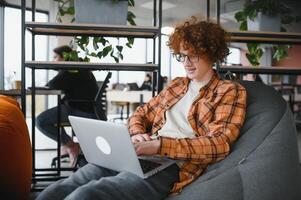 Portrait of Caucasian male freelancer in trendy apparel sitting at cafeteria table and doing remote work for programming design of public website, skilled software developer posing in coworking space. photo