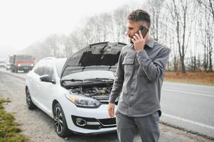 Sad driver calling car service, opening hood, having engine problem standing near broken car on the road. Car breakdown concept photo