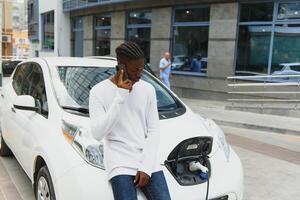 African man use smart phone while waiting and power supply connect to electric vehicles for charging the battery in car photo