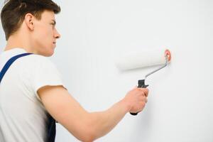 young man in blue overalls painting wall into white color with a roller photo