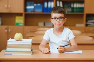 Concentrated schoolboy sitting at desk and writing in exercise book with classmate sitting behind photo