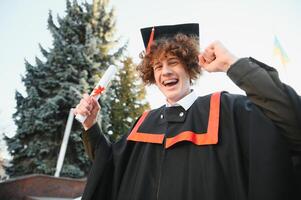 Happy smiling university graduate in mantle holding diploma in raised hand and expressing happiness over university building at background. Successful graduating from university or college photo