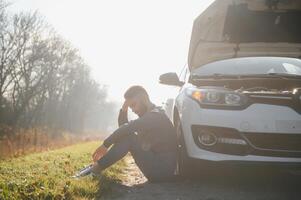 Picture of frustrated man sitting next to broken car with open hood photo