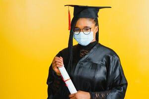 Portrait of a young african student in a graduate cap protective mask, on a yellow background, Graduation 2021 photo