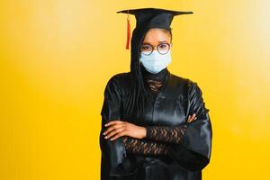 Portrait of a young african student in a graduate cap protective mask, on a yellow background, Graduation 2021 photo