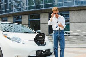 joven hermoso hombre participación cargando cable a eléctrico cargando estación punto en pie cerca su nuevo coche , mirando satisfecho. foto