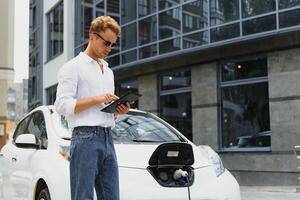 Business man standing near charging electric car and using tablet in the street. photo
