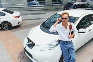 hombre cargando su lujo eléctrico coche a al aire libre estación en frente de moderno nuevo ciudad edificios foto