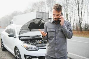 Sad driver calling car service, opening hood, having engine problem standing near broken car on the road. Car breakdown concept photo