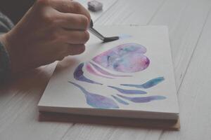 Woman paints in a sketchbook. Tools for painting on the white wooden desk. Painter work. photo