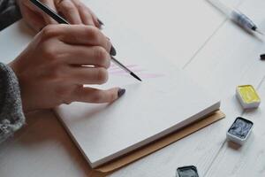 Woman paints in a sketchbook. Tools for painting on the white wooden desk. Painter work. photo