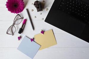 computadora portátil, vistoso memorándum hojas, los anteojos, aguja, rosado gerbera y elementos de decoración en un blanco de madera trabajo escritorio. hermosa hembra lugar de trabajo parte superior vista. foto