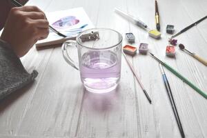 Woman paints in a sketchbook. Tools for painting on the white wooden desk. Painter work. photo