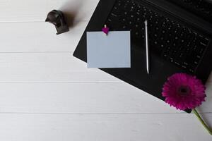 Black laptop keyboard, blue empty paper and purple gerbera on a white wooden work desk. photo