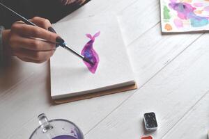 Woman paints in a sketchbook. Tools for painting on the white wooden desk. Painter work. photo