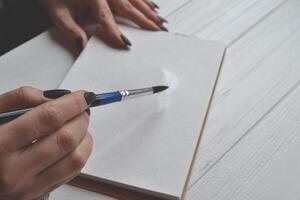 Woman paints in a sketchbook. Tools for painting on the white wooden desk. Painter work. photo