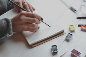 Woman paints in a sketchbook. Tools for painting on the white wooden desk. Painter work. photo