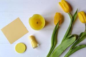 Yellow paper, tulips, candle and female accessories on a desk. Stylish background with place for text. Spring mood card. Yellow still life. photo