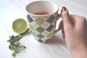 Woman's hand with a cup of natural tea. Lime and mint on the white wooden table. photo