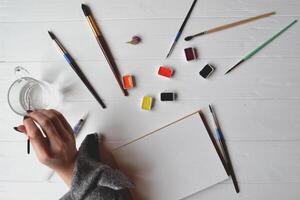 Woman paints in a sketchbook. Tools for painting on the white wooden desk. Painter work. photo