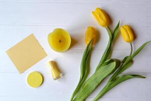 Yellow paper, tulips, candle and female accessories on a desk. Stylish background with place for text. Spring mood card. Yellow still life. photo