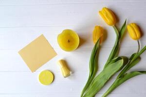 Yellow paper, tulips, candle and female accessories on a desk. Stylish background with place for text. Spring mood card. Yellow still life. photo