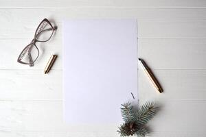 The sheet of white paper with pen, eyewear and branch of evergreen on the wooden desk. photo