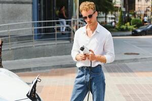 Stylish man with coffe cup in hand inserts plug into the electric car charging socket photo