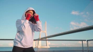 un mujer en un capucha y rojo guantes es practicando boxeo en un puente video