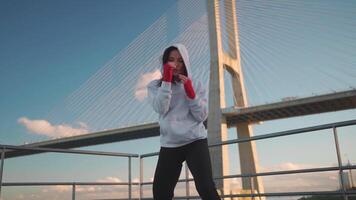 a woman in a hoodie and red gloves is practicing boxing on a bridge video