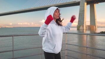 a woman in a hoodie and red gloves is practicing boxing on a bridge video