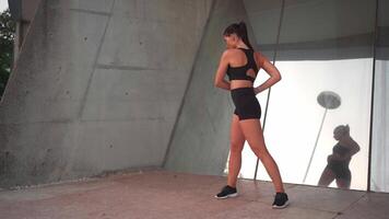 une femme dans une noir exercice costume Faire chaud en haut des exercices près miroir mur en plein air ville rue video