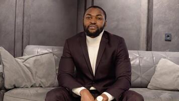 A young elegant bearded man in a classic wine suit is sitting on a sofa in a showroom or boutique video