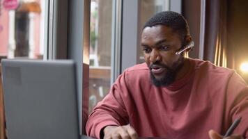 African american young man wear headset communicating by conference call speak looking at computer in cafe. Video chat job interview or distance language course class concept