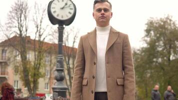 portrait de une Beau à la mode homme dans marron manteau contre le Contexte de rue horloges. Nouveau collection de à la mode vêtements video