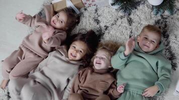 cuatro dulce niños laico abajo jn un alfombra cerca Navidad árbol y ondulación su piernas y manos. ver desde parte superior video