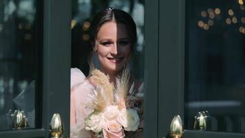 radiante novia por el ventana, un sonriente novia participación un ramo de flores poses por el ventana, su alegría iluminado por un destello de luz de sol. video