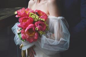 un hermosa Boda ramo de flores con rosado peonías en el manos de un novia en un blanco vestir con hinchado mangas foto
