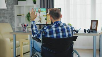 Freelancer in wheelchair waving during a business video call while working from home office.